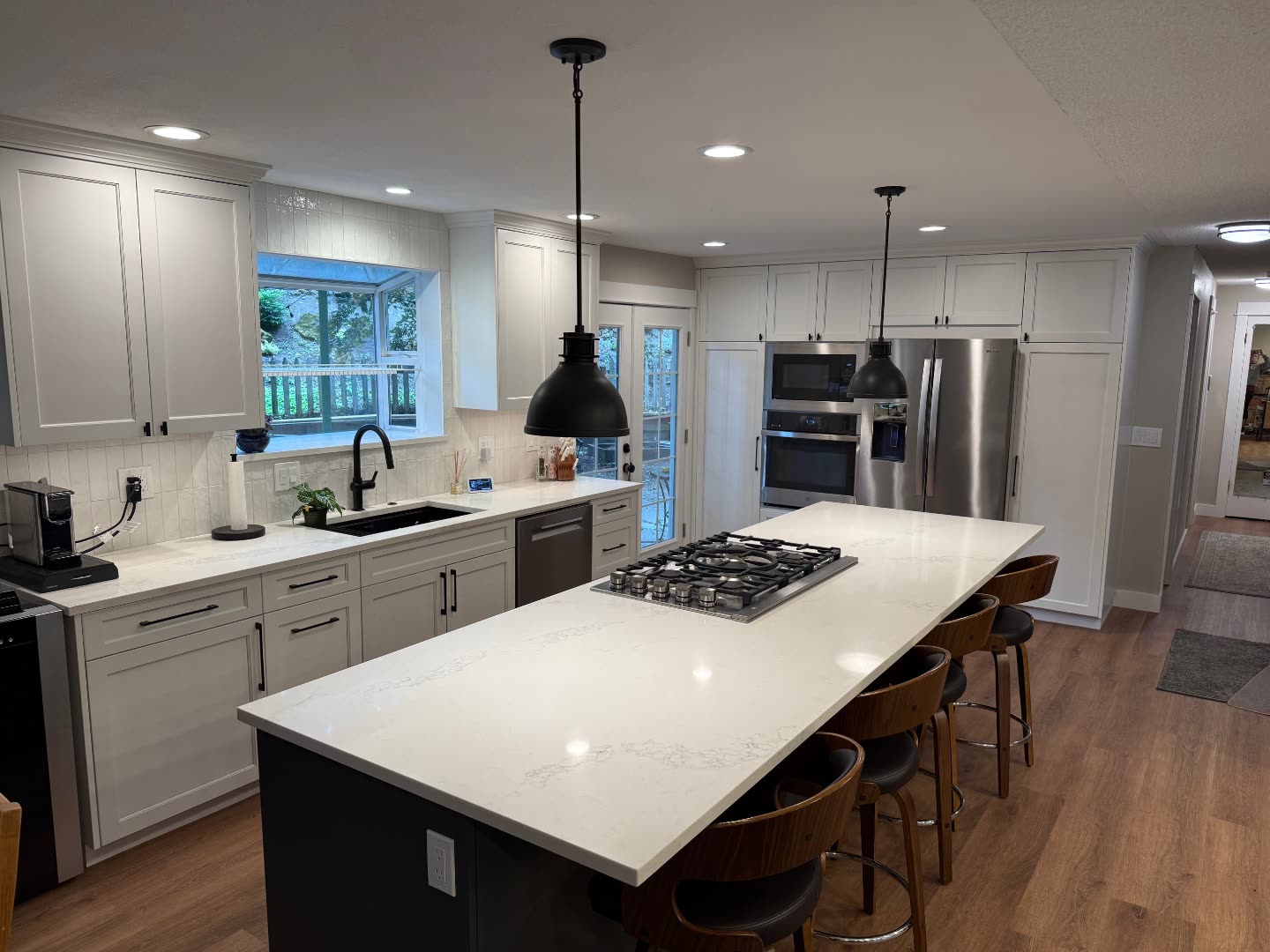 Jones residence kitchen after renovation — wide L-shape kitchen with white shaker cabinets, marble counters, charcoal island with gas cooktop and black industrial pendants.