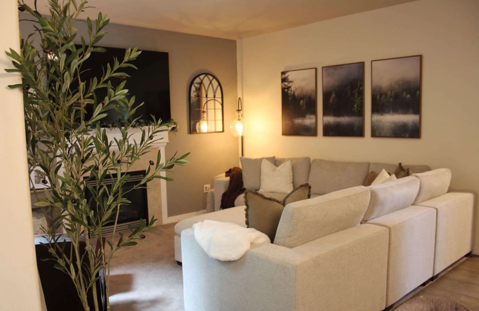 Modern living room with olive tree, sectional, and moody black-and-white triptych above fireplace at Lydon Residence.