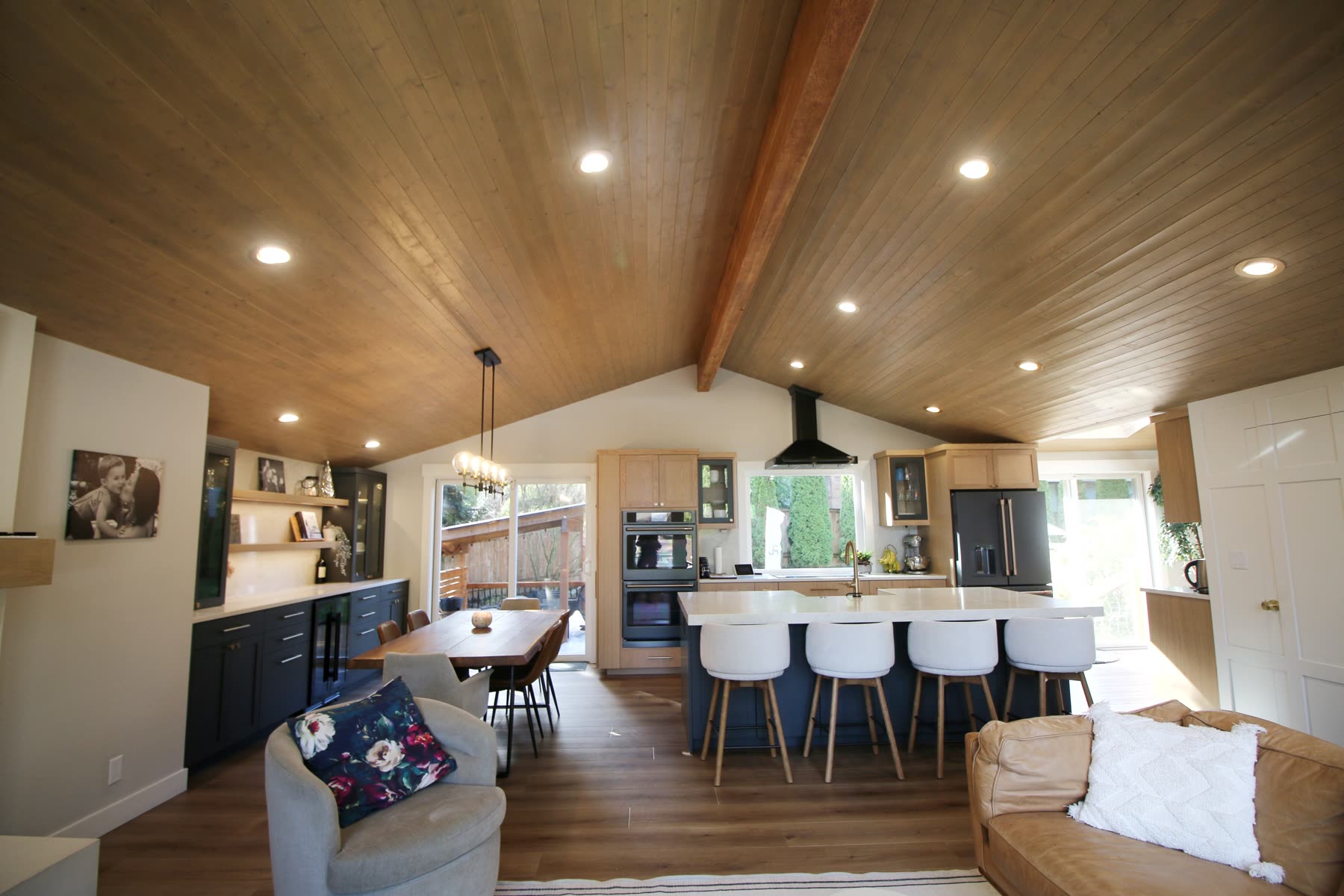Van Buuren residence — open great room with vaulted wood beams, oak and navy kitchen, live-edge dining table, and brass globe pendants.